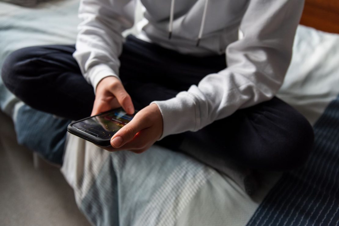 close up of a teenage boy sitting cross legged on his bed looking at his phone