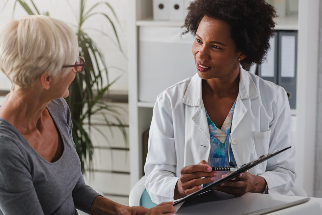 A female doctor sits at her desk and chats to an elderly female